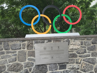 The image features five interlocking colored rings representing the Olympic symbol, mounted on a stone wall. A plaque with text is attached to the wall below the rings. Green foliage is visible in the background.