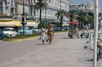 Two women in fashionable mid-20th century dresses are walking along a promenade lined with palm trees. Vintage cars are parked along the street, and there are several small kiosks and shops with outdoor seating. The architecture reflects a classic European style, with ornate details on the buildings. A vendor stall selling postcards and magazines is visible.