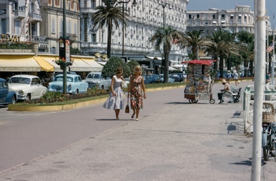 Two women in fashionable mid-20th century dresses are walking along a promenade lined with palm trees. Vintage cars are parked along the street, and there are several small kiosks and shops with outdoor seating. The architecture reflects a classic European style, with ornate details on the buildings. A vendor stall selling postcards and magazines is visible.