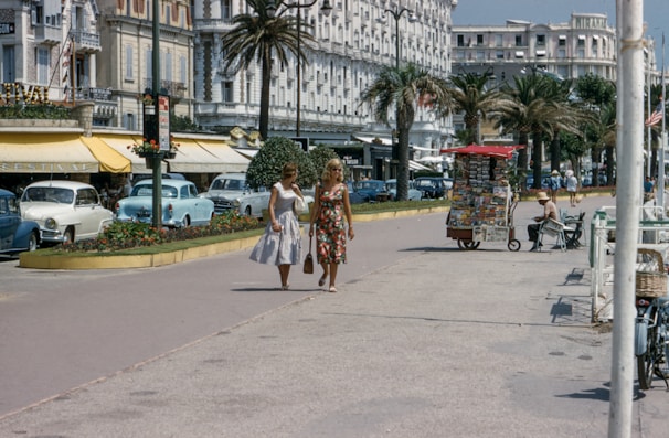Two women in fashionable mid-20th century dresses are walking along a promenade lined with palm trees. Vintage cars are parked along the street, and there are several small kiosks and shops with outdoor seating. The architecture reflects a classic European style, with ornate details on the buildings. A vendor stall selling postcards and magazines is visible.