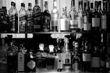 A collection of various bottles arranged on two shelves. The upper shelf contains different brands of whiskey, while the lower shelf has an assortment of spirits, including gin and port. The scene is captured in black and white, emphasizing the shapes and labels of the bottles.