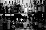 A collection of various bottles arranged on two shelves. The upper shelf contains different brands of whiskey, while the lower shelf has an assortment of spirits, including gin and port. The scene is captured in black and white, emphasizing the shapes and labels of the bottles.