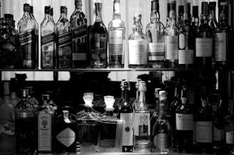 A collection of various bottles arranged on two shelves. The upper shelf contains different brands of whiskey, while the lower shelf has an assortment of spirits, including gin and port. The scene is captured in black and white, emphasizing the shapes and labels of the bottles.