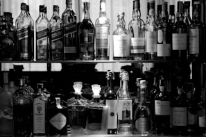 A collection of various bottles arranged on two shelves. The upper shelf contains different brands of whiskey, while the lower shelf has an assortment of spirits, including gin and port. The scene is captured in black and white, emphasizing the shapes and labels of the bottles.