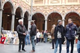 A group of people are gathered in a historical courtyard with ornate architectural details, including arches and sculptures. Several individuals are standing and conversing, while others are browsing through artwork displayed on easels. The atmosphere appears lively and social.