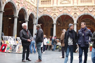 A group of people are gathered in a historical courtyard with ornate architectural details, including arches and sculptures. Several individuals are standing and conversing, while others are browsing through artwork displayed on easels. The atmosphere appears lively and social.