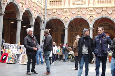 A group of people are gathered in a historical courtyard with ornate architectural details, including arches and sculptures. Several individuals are standing and conversing, while others are browsing through artwork displayed on easels. The atmosphere appears lively and social.
