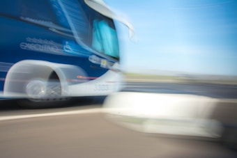 A moving coach bus captured in motion blur, creating a dynamic and fast-paced effect. The bus is predominantly blue and white, featuring a logo and text on its side. The background shows a road and sky blurred due to the motion.
