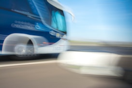 A moving coach bus captured in motion blur, creating a dynamic and fast-paced effect. The bus is predominantly blue and white, featuring a logo and text on its side. The background shows a road and sky blurred due to the motion.