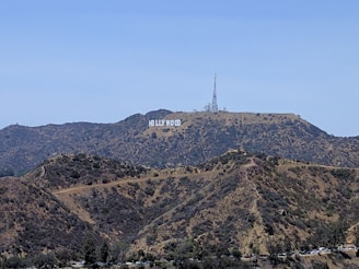 A large white sign reading 'Hollywood' is displayed on a hillside surrounded by rugged terrain with sparse vegetation. A tall communication tower and other structures are visible on the peak.