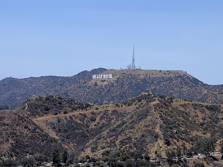 A large white sign reading 'Hollywood' is displayed on a hillside surrounded by rugged terrain with sparse vegetation. A tall communication tower and other structures are visible on the peak.