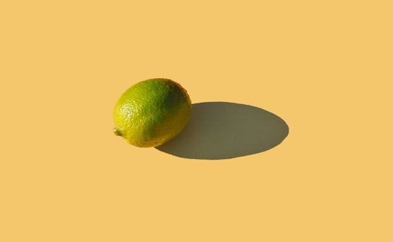A single lime is placed on a solid pastel yellow background, casting a defined shadow. The lime's texture is bumpy and shiny, indicating its freshness, and its vibrant green color contrasts with the smooth background.