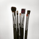 A collection of paintbrushes with metallic handles and bristles of various lengths and shapes, standing upright against a plain background.