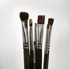 A collection of paintbrushes with metallic handles and bristles of various lengths and shapes, standing upright against a plain background.