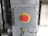 A metallic panel with a prominent red emergency stop button is mounted on an industrial setting. The button is round with a yellow background and labeled 'EMERGENCY STOP'. It is surrounded by cables and metallic components, suggesting its placement in a machinery or factory environment.