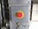 A metallic panel with a prominent red emergency stop button is mounted on an industrial setting. The button is round with a yellow background and labeled 'EMERGENCY STOP'. It is surrounded by cables and metallic components, suggesting its placement in a machinery or factory environment.