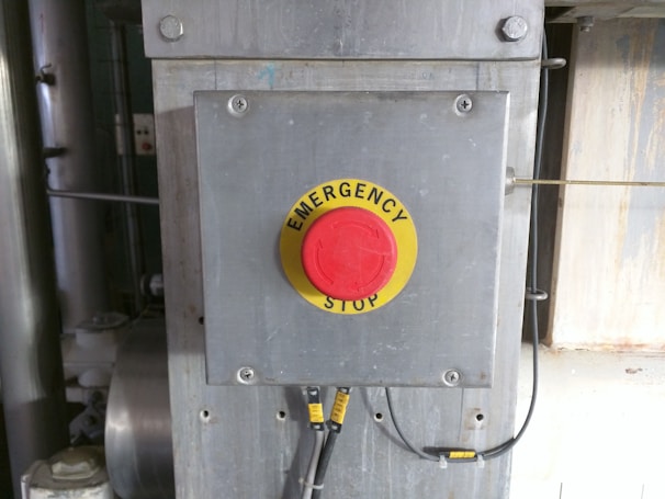 A metallic panel with a prominent red emergency stop button is mounted on an industrial setting. The button is round with a yellow background and labeled 'EMERGENCY STOP'. It is surrounded by cables and metallic components, suggesting its placement in a machinery or factory environment.