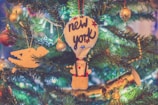 A close-up of a Christmas tree decorated with various ornaments. Prominently displayed is a hot air balloon ornament made of felt with 'new york' written on it in black script and adorned with star decorations. Below the balloon, a small Santa figure peeks out. Other ornaments visible include a wooden bird and gold baubles, with strings of beads wrapping around the branches. The lights of the tree create a warm, festive glow.