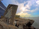 A coastal scene with a stone wall featuring a painted image of a young girl in a white dress and a red rose on her chest. The background displays a cloudy sky, the ocean, and a distant cityscape. The foreground includes rocky and sandy terrain.