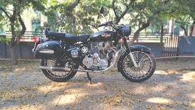 A classic motorcycle is parked on a pavement against a backdrop of trees and a fence. The bike has a polished black finish with chrome detailing. Fallen leaves are scattered on the ground, adding a touch of autumn to the scene.