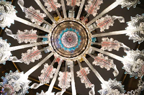 Intricately designed, the image features a symmetrical chandelier viewed from below. The chandelier has ornate glass elements radiating outward, with highlights of pink, blue, and clear glass. The craftsmanship showcases detailed patterns resembling feathers or leaves on the arms extending from the center.