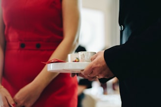 A person in a red dress stands beside another individual in a dark suit jacket holding a white tray with a cup and a small packet. The setting suggests a formal or social gathering.