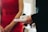 A person in a red dress stands beside another individual in a dark suit jacket holding a white tray with a cup and a small packet. The setting suggests a formal or social gathering.