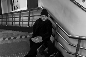 An elderly person wearing a beanie and scarf sits on a stairway, holding a folded item. The setting is indoors with metal railings and a poster on the wall.
