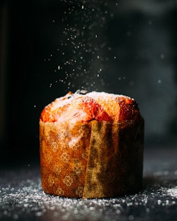 A freshly baked panettone dusted with powdered sugar sits on a dark surface. The pastry is enveloped in a decorative brown paper with a subtle floral pattern. Powdered sugar is being sprinkled from above, creating a soft snowfall effect.