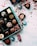 An assortment of decorative chocolate pieces placed in a blue box, with colorful wrapping scattered around. Red strings and ribbons are artistically strewn across a white surface dusted with cocoa powder.