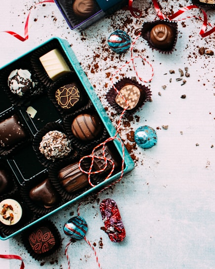 An assortment of decorative chocolate pieces placed in a blue box, with colorful wrapping scattered around. Red strings and ribbons are artistically strewn across a white surface dusted with cocoa powder.