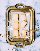 An ornate, vintage-style metal tray holds six rectangular shortbread cookies, arranged on a piece of parchment paper. Around these cookies are decorative paper snowflakes and golden curling ribbon. The tray is set on a marbled surface.