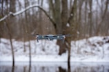 A drone hovering in the air during a snowy day in a forested area. Snowflakes are visible in the air, and bare trees can be seen in the background, along with a snow-covered ground.
