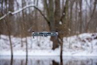 A drone hovering in the air during a snowy day in a forested area. Snowflakes are visible in the air, and bare trees can be seen in the background, along with a snow-covered ground.