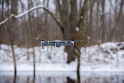 A drone hovering in the air during a snowy day in a forested area. Snowflakes are visible in the air, and bare trees can be seen in the background, along with a snow-covered ground.