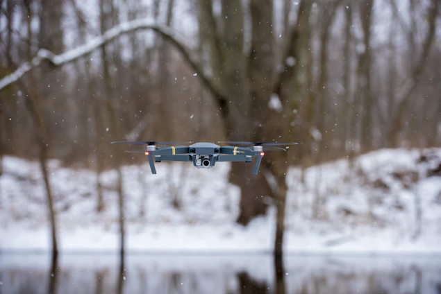 A drone hovering in the air during a snowy day in a forested area. Snowflakes are visible in the air, and bare trees can be seen in the background, along with a snow-covered ground.