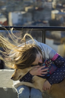 A person is embracing a dog with affection, holding the animal close while their hair flows in the wind. The background shows a blurred view of buildings and the cityscape under soft sunlight.