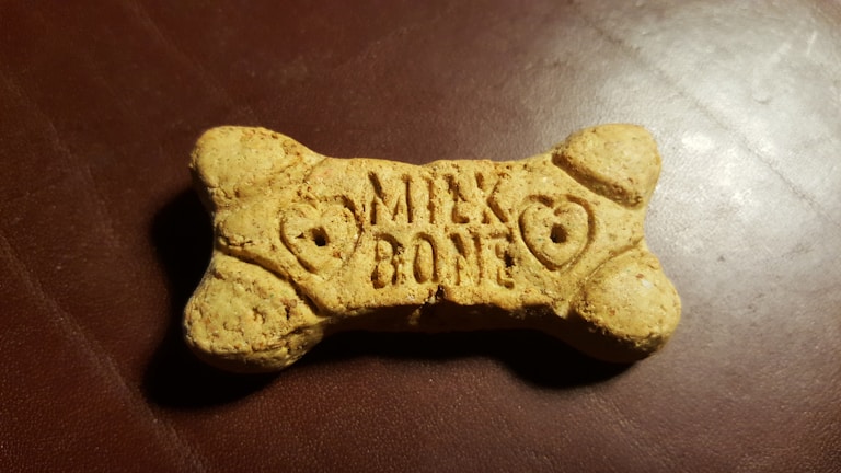 A dog biscuit shaped like a bone with the words 'Milk Bone' engraved on its surface, placed on a textured, dark brown background.
