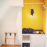 A small, open wardrobe area with a bright yellow painted interior. On the right, there are wooden hangers on a rod, with a single dark garment hanging. The middle shelf holds two blue helmets and other small items. On the left, a white robe hangs beside a wooden folding luggage rack.