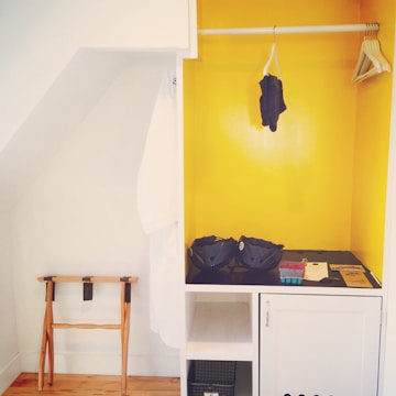 A small, open wardrobe area with a bright yellow painted interior. On the right, there are wooden hangers on a rod, with a single dark garment hanging. The middle shelf holds two blue helmets and other small items. On the left, a white robe hangs beside a wooden folding luggage rack.