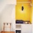 A small, open wardrobe area with a bright yellow painted interior. On the right, there are wooden hangers on a rod, with a single dark garment hanging. The middle shelf holds two blue helmets and other small items. On the left, a white robe hangs beside a wooden folding luggage rack.