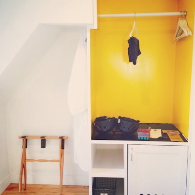 A small, open wardrobe area with a bright yellow painted interior. On the right, there are wooden hangers on a rod, with a single dark garment hanging. The middle shelf holds two blue helmets and other small items. On the left, a white robe hangs beside a wooden folding luggage rack.