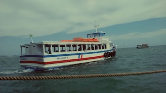 A ferry boat is navigating through a body of water with its hull painted in blue and white with a red stripe. The green and yellow flag on the stern flutters in the breeze. Several passengers are visible through the windows. The sky is partly cloudy, and the water is slightly choppy. Another boat can be seen in the distance.