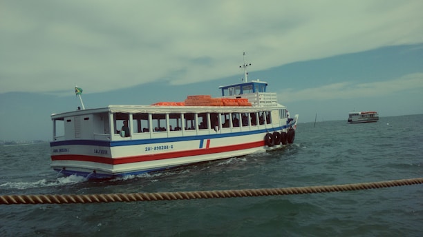 A ferry boat is navigating through a body of water with its hull painted in blue and white with a red stripe. The green and yellow flag on the stern flutters in the breeze. Several passengers are visible through the windows. The sky is partly cloudy, and the water is slightly choppy. Another boat can be seen in the distance.