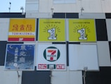 Several business signs, including a Torikizoku sign on a yellow and red background, a karaoke specialty store sign with a bright yellow background, and a 7-Eleven convenience store logo. The signs are mounted on a building exterior, featuring lighting fixtures below each sign.