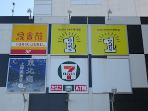 Several business signs, including a Torikizoku sign on a yellow and red background, a karaoke specialty store sign with a bright yellow background, and a 7-Eleven convenience store logo. The signs are mounted on a building exterior, featuring lighting fixtures below each sign.