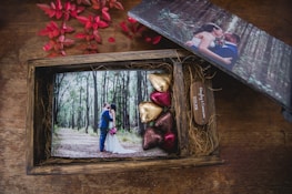 A wooden box containing a wedding photograph, a small wooden plaque with names and a date, and several heart-shaped chocolates wrapped in colorful foils. The photograph shows a couple in wedding attire surrounded by trees in a forest setting. There are also red leaves nearby, adding to the aesthetic of the composition.