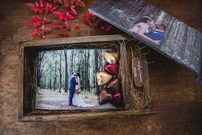 A wooden box containing a wedding photograph, a small wooden plaque with names and a date, and several heart-shaped chocolates wrapped in colorful foils. The photograph shows a couple in wedding attire surrounded by trees in a forest setting. There are also red leaves nearby, adding to the aesthetic of the composition.