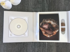 An open photo album with a CD holder on the left page and a photograph on the right. The photograph depicts a newborn baby wrapped in a brown blanket, lying on a fluffy, soft background. The album is placed on a dark wooden surface with two white orchids visible in the top left corner.