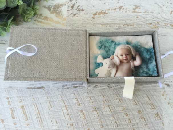 A textured box with a light brown fabric exterior is open to reveal a photo of a baby inside. The baby is smiling, wearing a white knitted hat, and cuddling a white stuffed animal against a fluffy teal backdrop. The box is placed on a rustic wooden surface and features a white ribbon on the side.